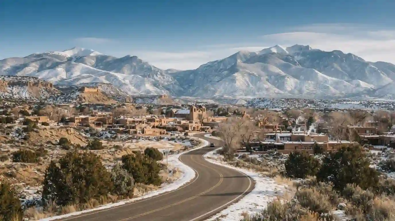 Snow-covered New Mexico mountains overlooking an adobe-style town and scenic winter road.